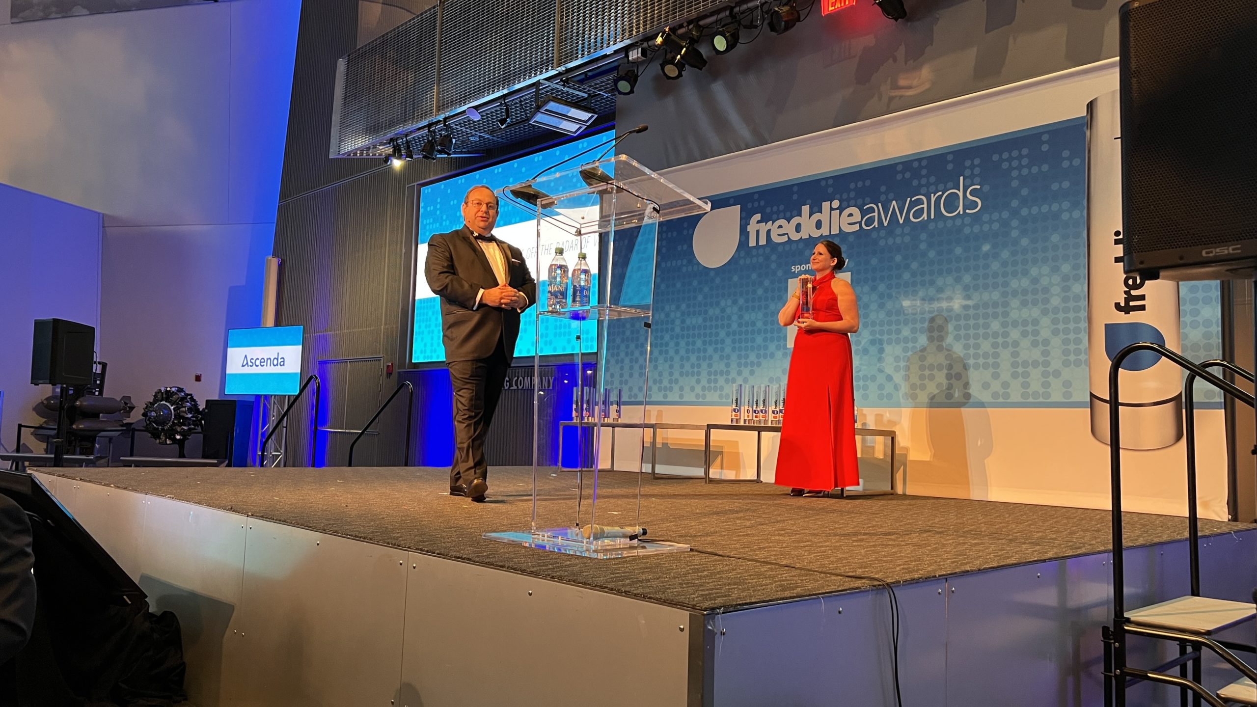 a man in a suit and tie standing on a stage with a podium with water bottles