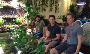 a group of men sitting on a bench in front of a pond with plants