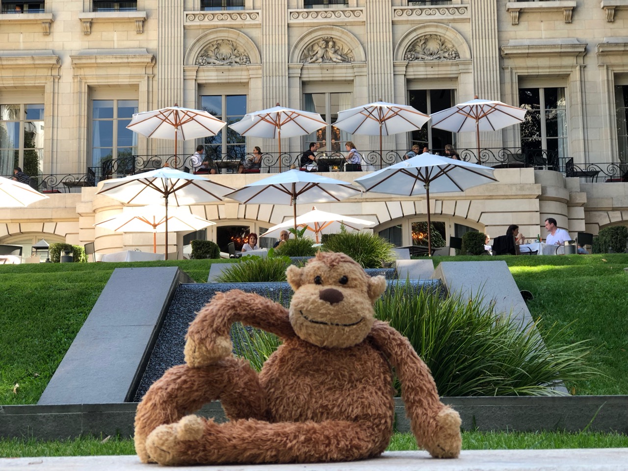 a stuffed monkey sitting on a stone ledge in front of a building with white umbrellas